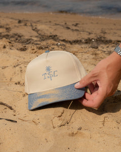 Beige cap with blue brim held by a hand on a sandy beach.